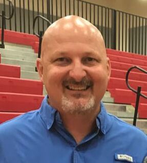Smiling man in a blue shirt stands in a gymnasium with red bleachers. He appears approachable and engaged, showcasing a community event atmosphere.