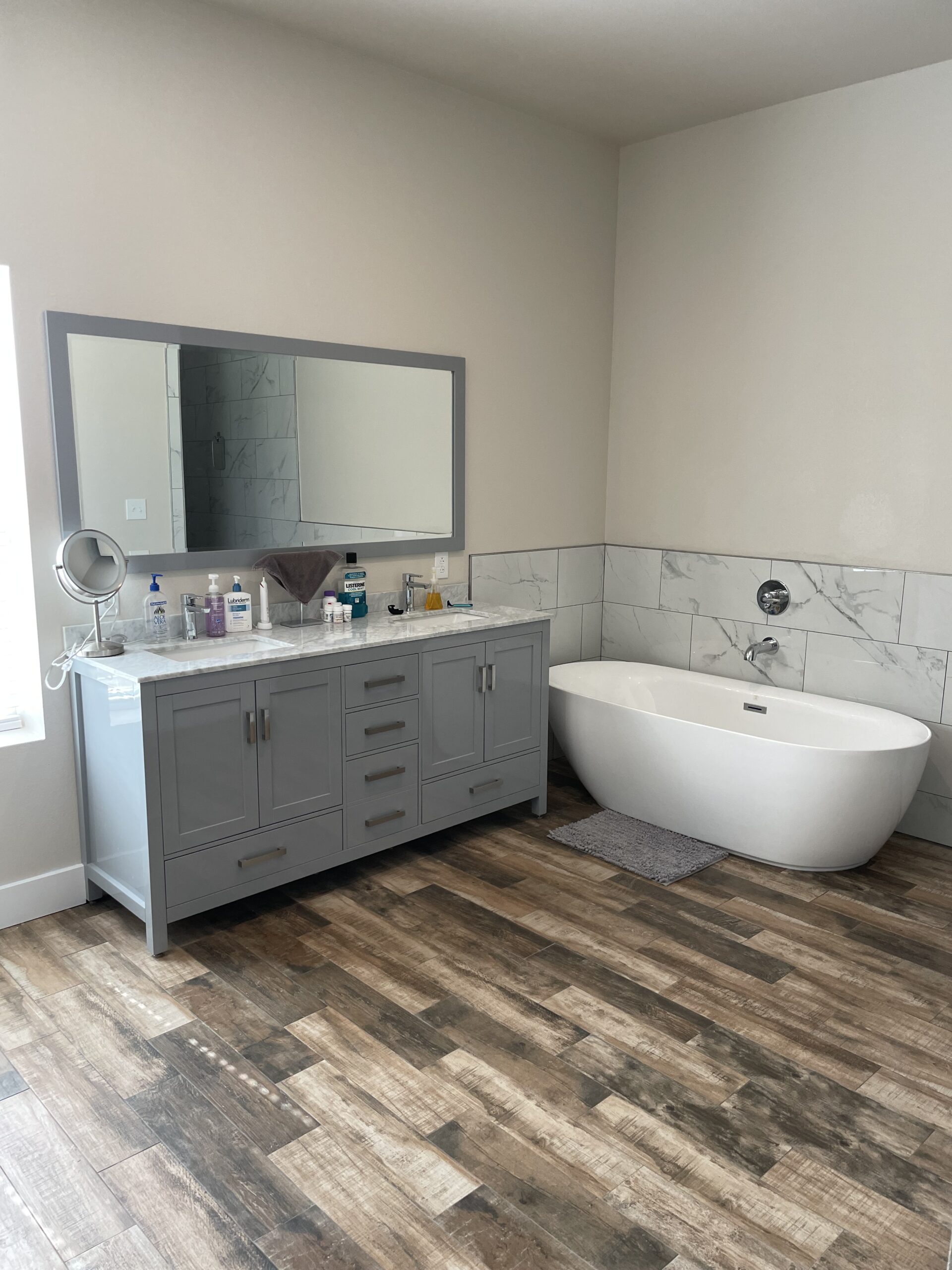 Modern bathroom featuring a gray vanity with a marble countertop, a large mirror, a freestanding bathtub, and stylish wood-like flooring, installed through our plumbing service in Georgetown, TX. Call today to bring your dream bathroom to life.