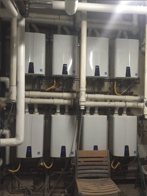 Eight white water heaters are mounted on a wall with pipes and yellow gas lines. A wooden chair is placed in front, indicating a maintenance area.