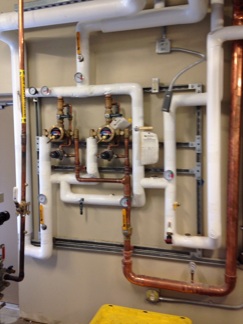 Complex plumbing system on a wall, featuring copper and PVC pipes, valves, and gauges, indicating water flow and pressure management.